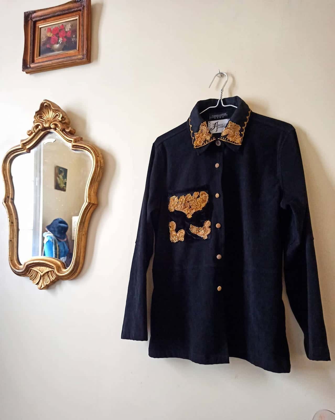 Chemise noire sur un cintre, col et poitrine brodés de motifs dorés, à côté d’un miroir doré