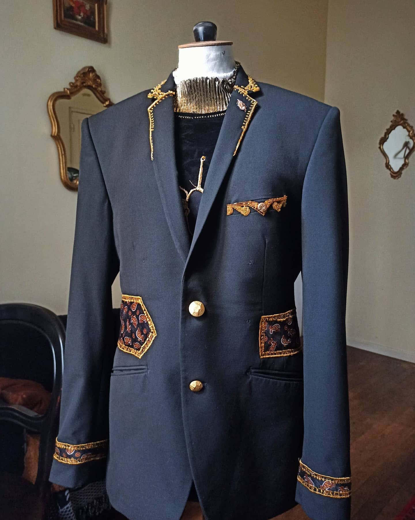 Blazer sombre sur un mannequin de couture, revers brodés de doré, poches et poignets à empiècements à motif cachemire, boutons dorés