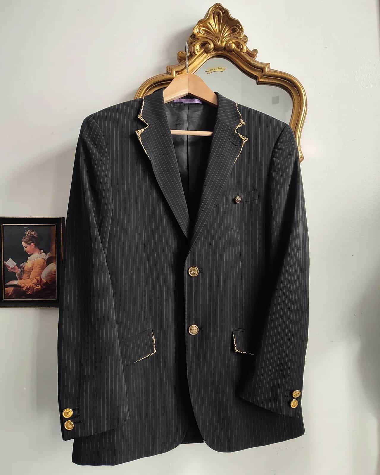 Blazer noir à fines rayures sur un cintre, devant un miroir doré : revers et rabats de poches bordés de broderie dorée, boutons dorés
