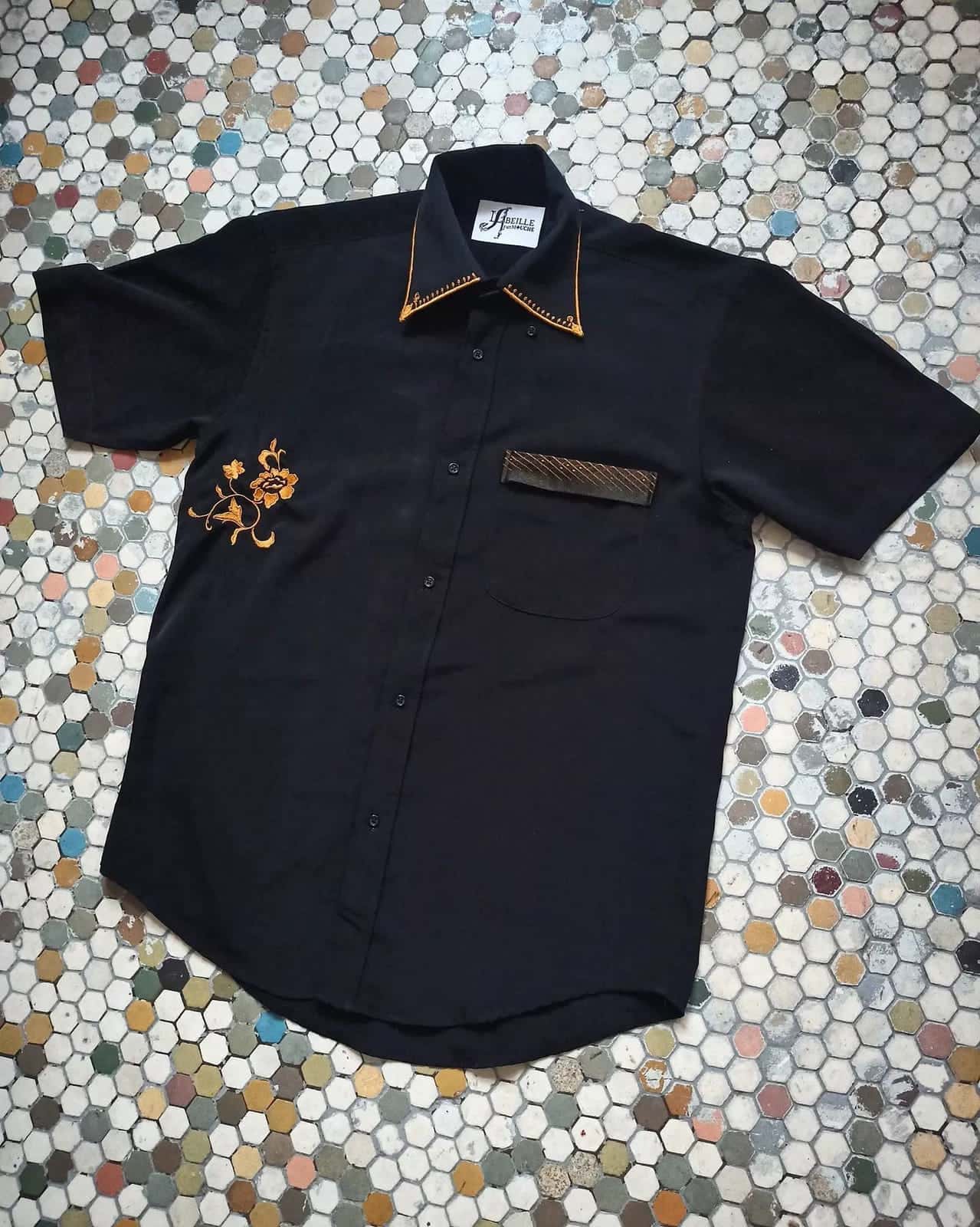 Chemise noire à manches courtes posée à plat, col bordé de doré, fleur brodée dorée sur le devant, poche à passepoil