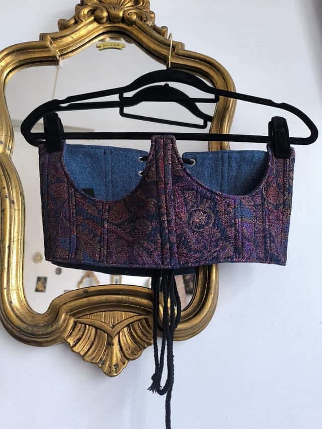 Corset à motif cachemire violet et empiècements en jean, sur un cintre devant un miroir doré