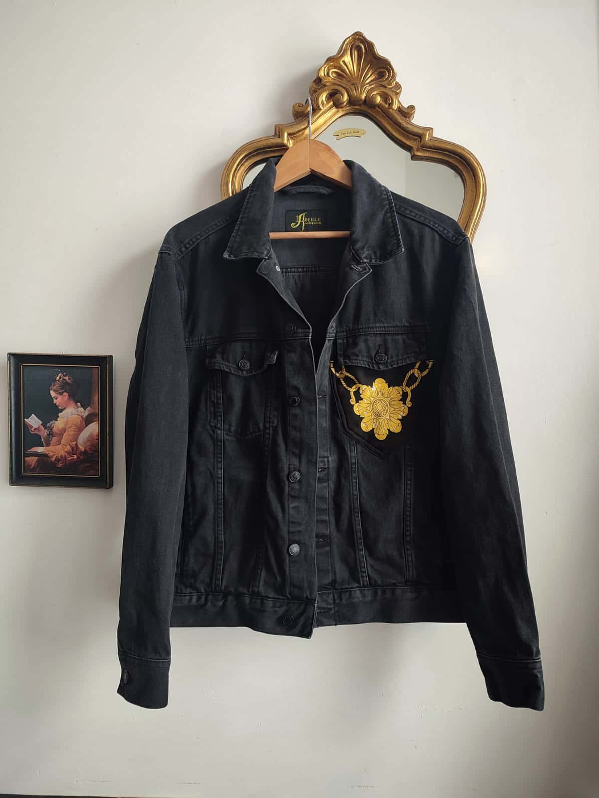 Veste en jean noire sur un cintre, devant un miroir doré : une fleur dorée et une chaîne dorée sur la poche poitrine