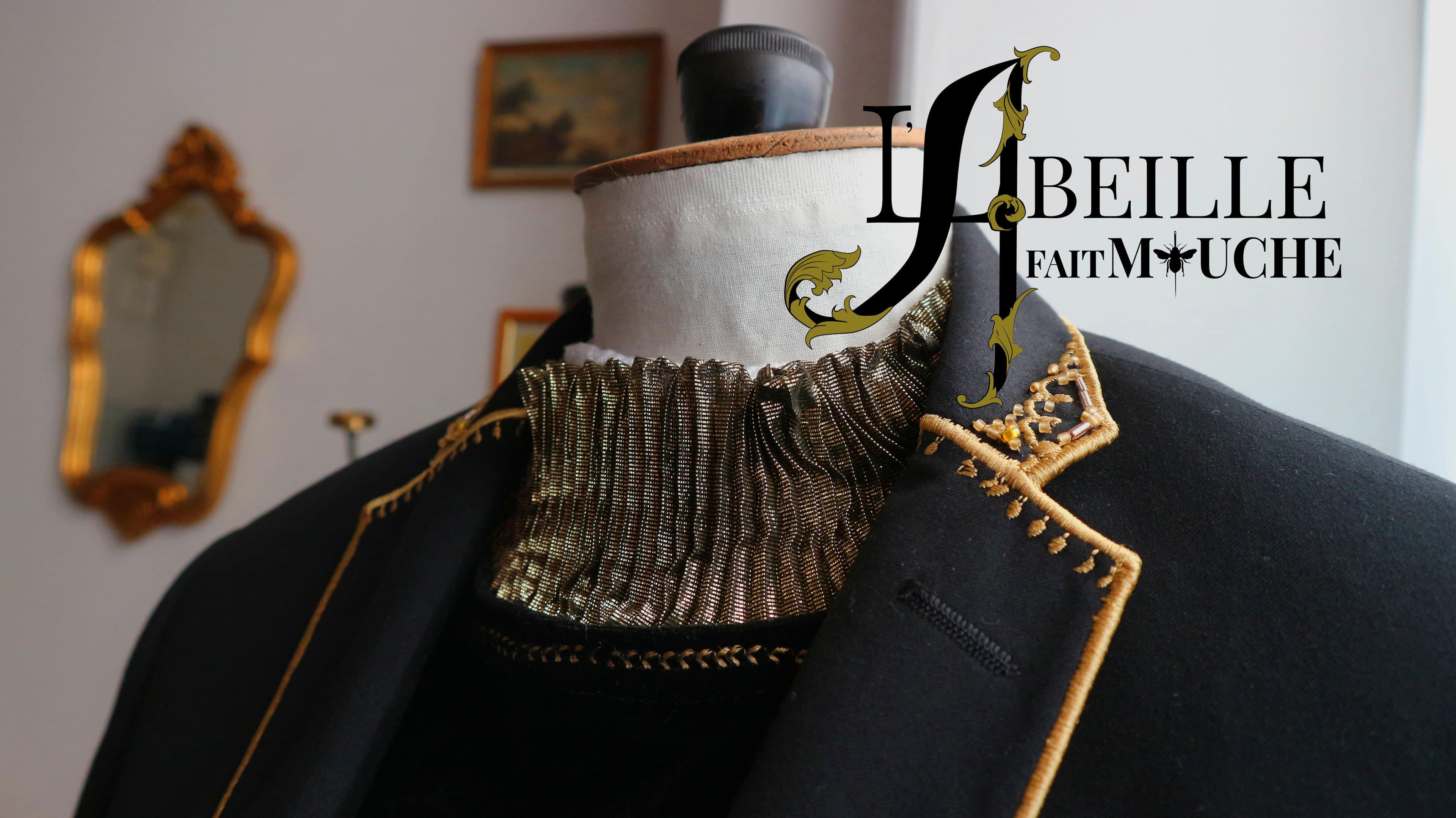 Veste noire sur un mannequin de couture, col montant plissé doré et revers bordé de broderie dorée, sous le logo de L’Abeille Fait Mouche. Un miroir doré au mur.