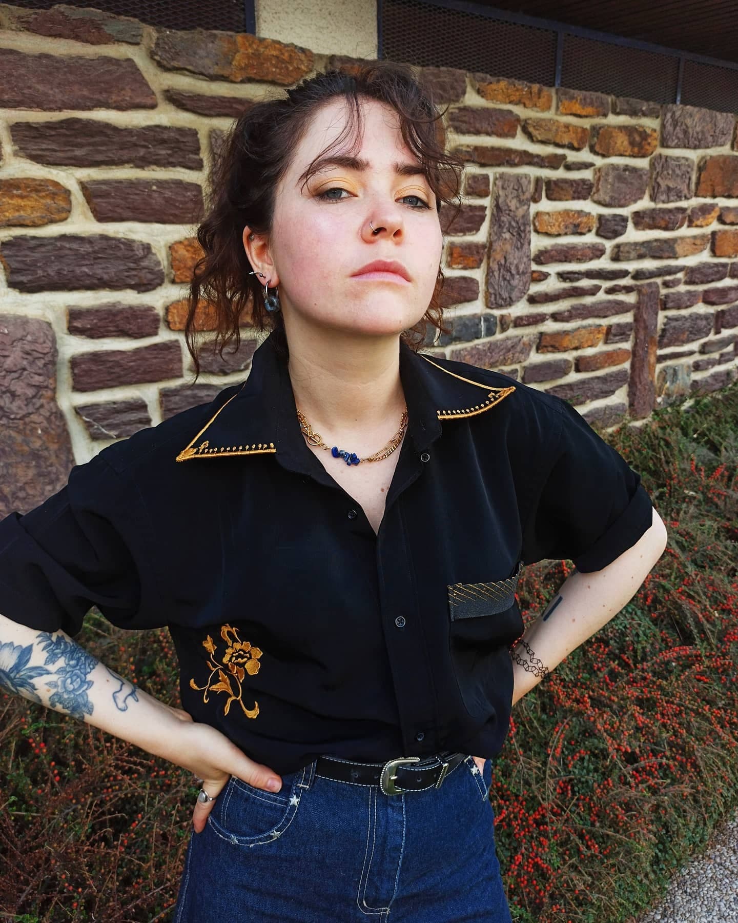 Chemise noire à manches courtes, col bordé de doré et fleur brodée dorée sur le devant, portée avec un jean bleu