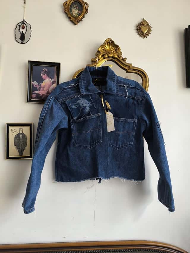 Veste en jean courte assemblée de plusieurs jeans, sur un cintre devant un miroir doré